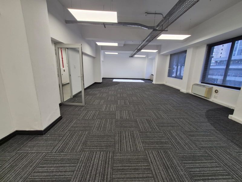 A spacious, open-plan office area featuring modern carpet tiles, suspended LED lighting, exposed cable management, and large windows providing ample natural light.