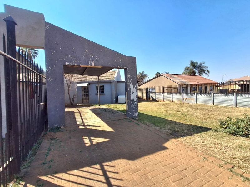 Front exterior view featuring a paved driveway, a carport structure with peeling paint, and metal perimeter fencing surrounding the property.