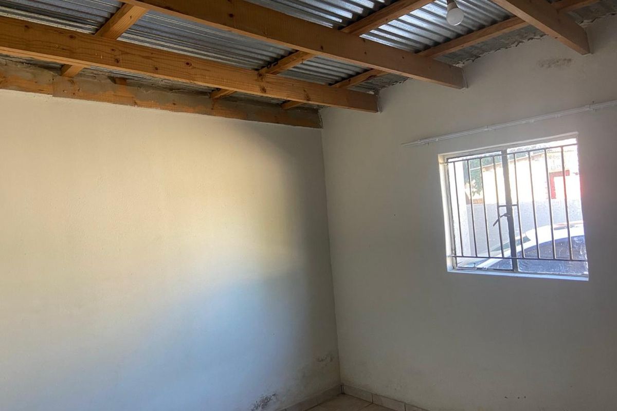 A basic, unfinished room featuring exposed timber roof beams, tiled flooring, and a window fitted with security bars.