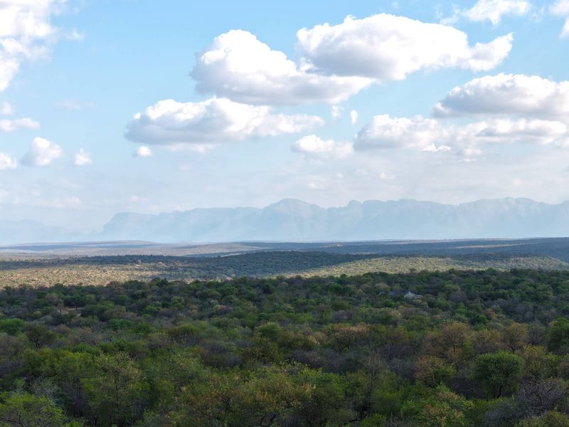 A scenic, elevated landscape view showcasing expansive natural bushveld and distant mountain ranges under a clear blue sky.