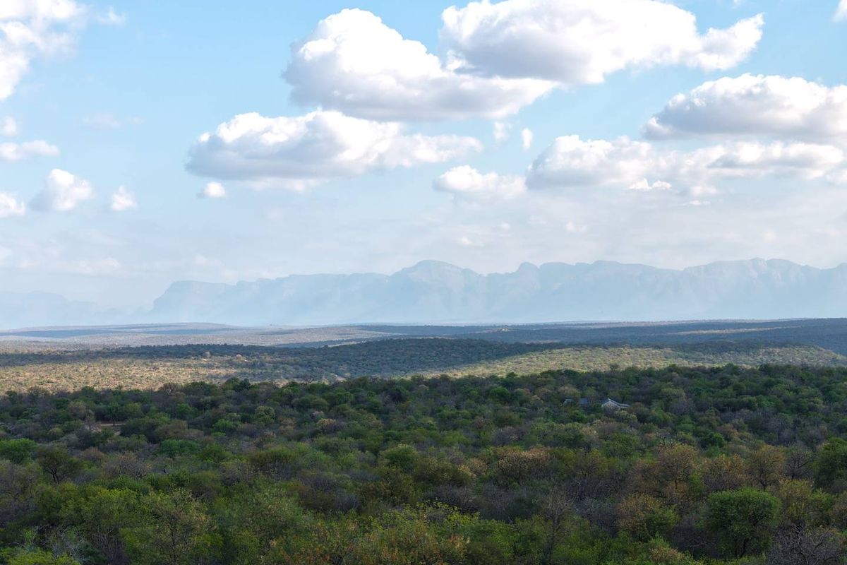 A scenic, elevated landscape view showcasing expansive natural bushveld and distant mountain ranges under a clear blue sky.