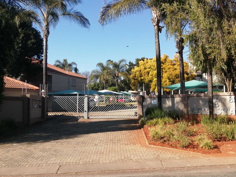 Front entrance view of the apartment complex featuring a paved driveway, secure metal gate, perimeter wall with electric fencing, and shaded parking bays.