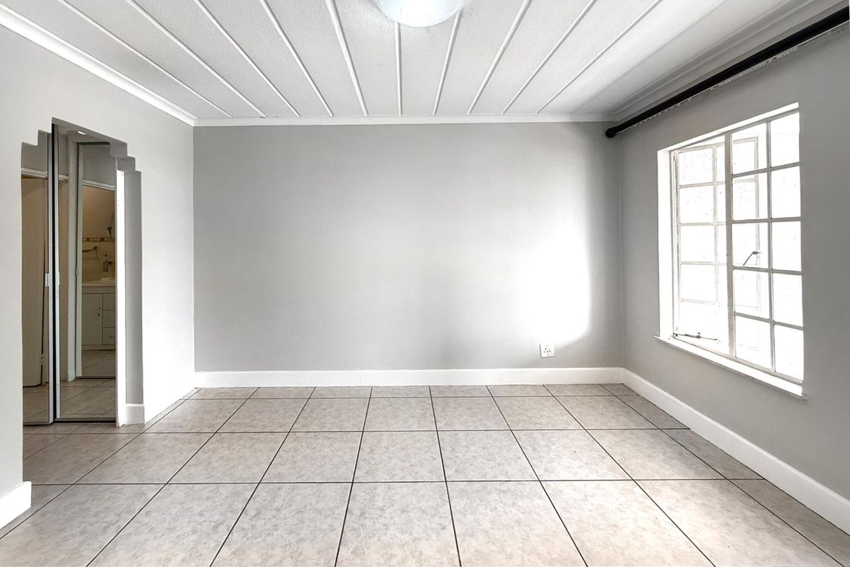 A bright, empty bedroom featuring tiled flooring, a paneled ceiling, and a window, with an adjacent doorway leading to a bathroom.