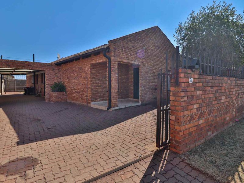 Front view of a facebrick house featuring a paved driveway, covered carport, and secure perimeter wall with a metal gate.