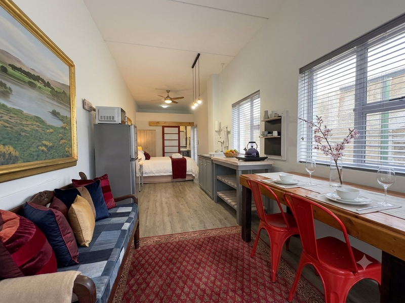 An open-plan cottage living space featuring laminate flooring, a compact kitchen area, and a bedroom zone with a ceiling fan and natural light.