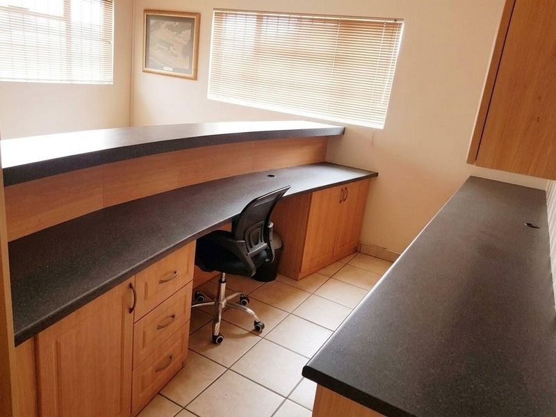 A functional office space featuring built-in laminate desks with dark countertops, tiled flooring, and fitted venetian blinds.