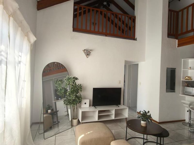 Spacious living area featuring high vaulted ceilings with exposed timber beams, a mezzanine level with timber balustrade, and bright tiled flooring.
