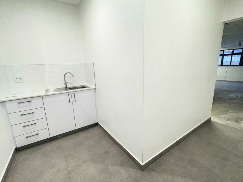 A clean, modern kitchenette area featuring white built-in cabinetry, a stainless steel sink, and neutral tiled flooring in an open office environment.
