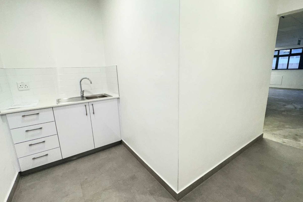 A clean, modern kitchenette area featuring white built-in cabinetry, a stainless steel sink, and neutral tiled flooring in an open office environment.