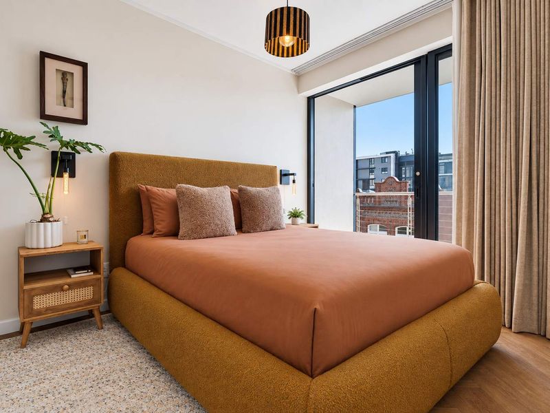 A well-maintained bedroom featuring hardwood flooring, modern wall-mounted lighting, and a large sliding glass door providing access to a private balcony.