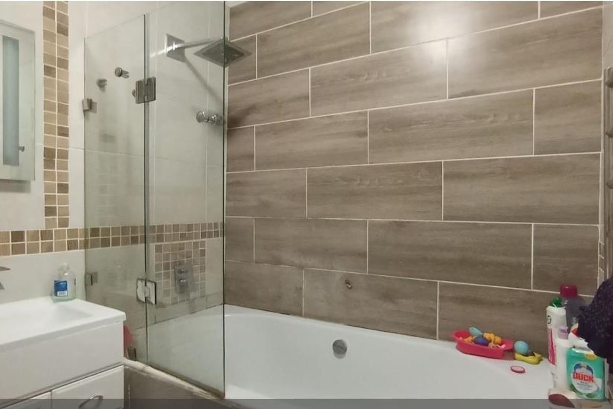 Modern bathroom featuring a bathtub with a glass shower screen, rainfall shower head, vanity basin, and heated towel rail with wood-look wall tiling.