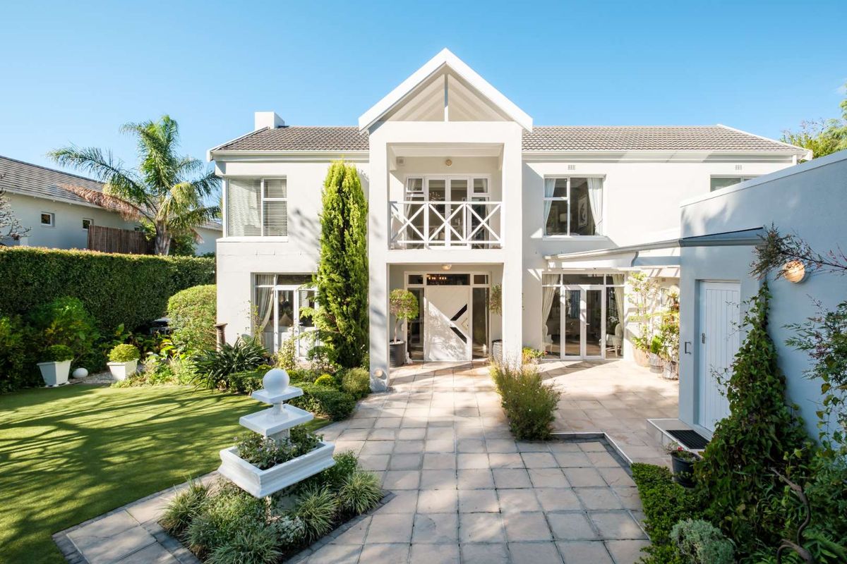A well-maintained rear exterior view featuring a paved patio, landscaped garden, central fountain, and a two-story facade with a balcony and french doors.