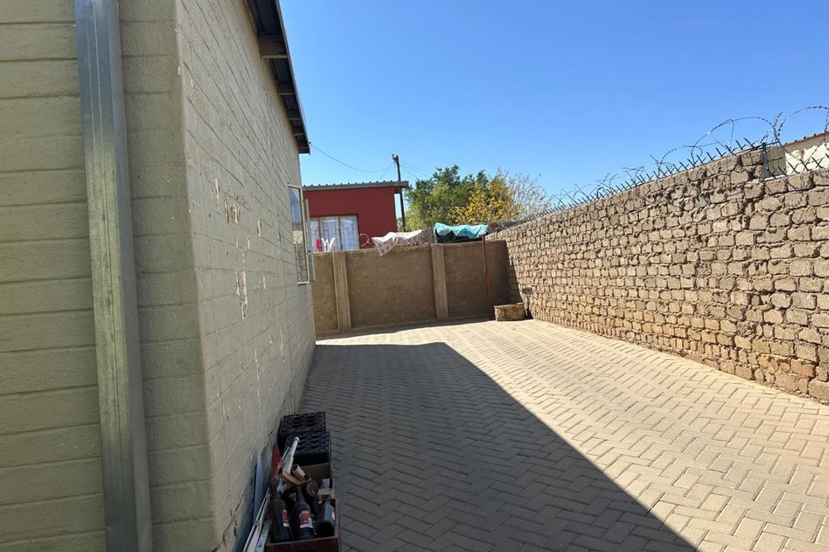 A rear exterior view showing a paved side yard, brick boundary wall with security razor wire, and the side elevation of the house.