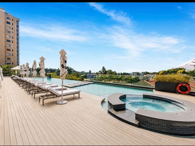 A modern rooftop pool area featuring an infinity edge swimming pool, integrated spa, composite decking, and glass balustrades with expansive views.