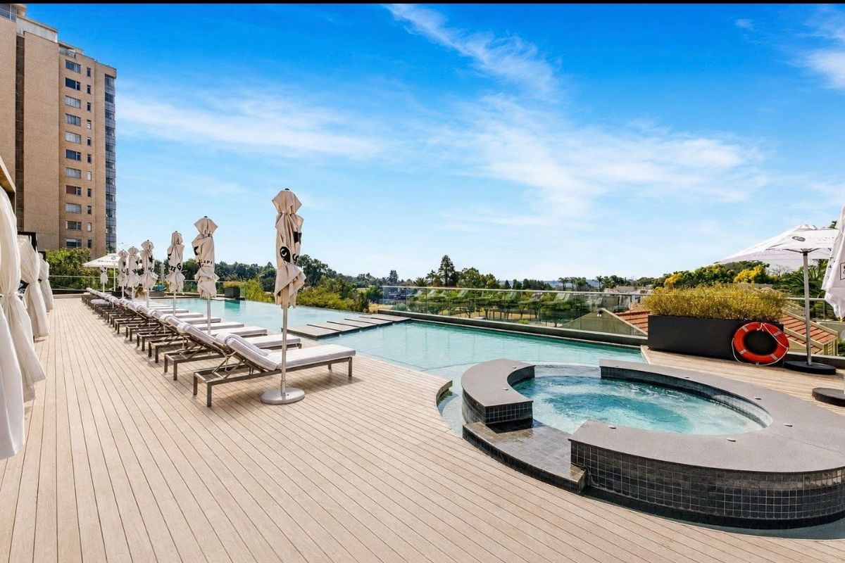 A modern rooftop pool area featuring an infinity edge swimming pool, integrated spa, composite decking, and glass balustrades with expansive views.