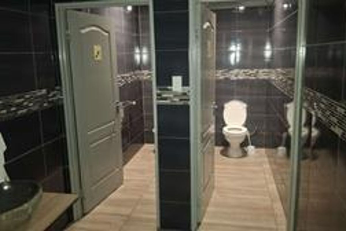 A functional office restroom featuring dark wall tiling, wood-look floor tiles, dual toilet cubicles, and a modern vessel sink vanity.