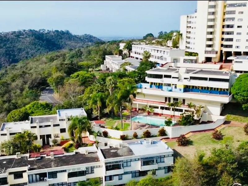 Aerial view of a multi-level apartment complex featuring landscaped grounds, a swimming pool, and scenic hillside surroundings.
