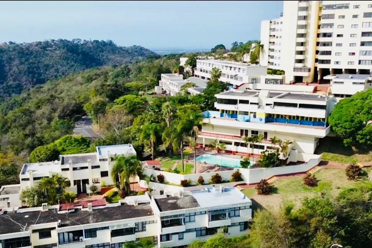 Aerial view of a multi-level apartment complex featuring landscaped grounds, a swimming pool, and scenic hillside surroundings.