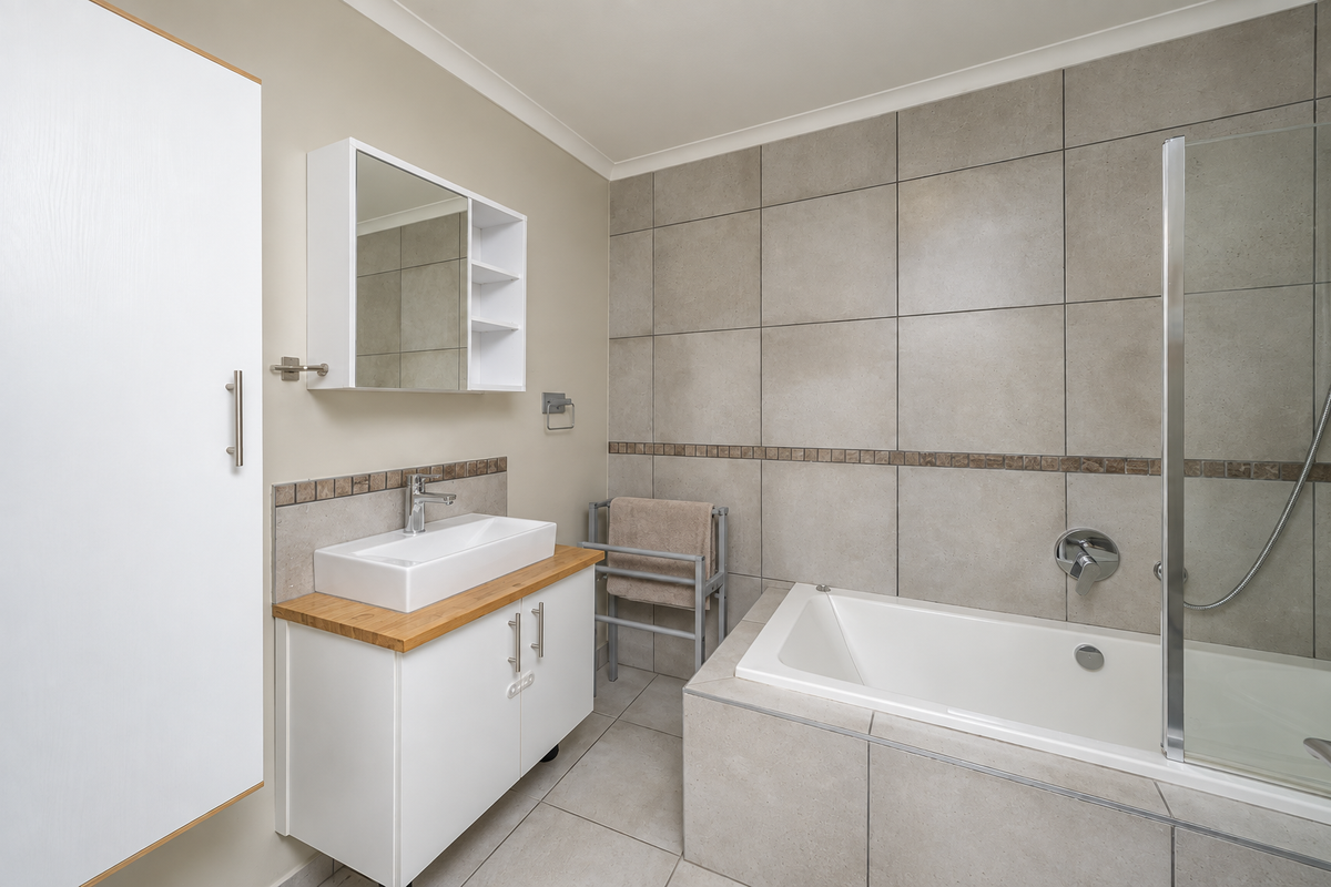 A modern, well-maintained bathroom featuring a vessel sink vanity, built-in bathtub with a glass shower screen, and neutral large-format wall and floor tiles.