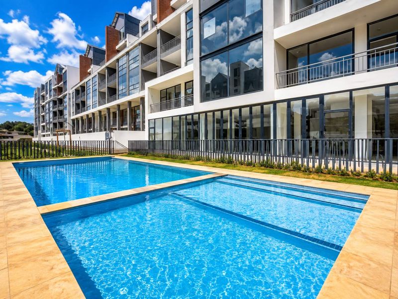 A well-maintained swimming pool area featuring a tiled surround and metal fencing, set against a modern multi-story apartment building with glass balconies.