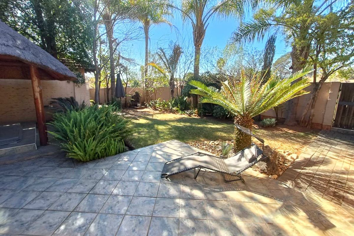 A private rear garden featuring a tiled patio area, established palm trees, lush greenery, and a thatched lapa structure.