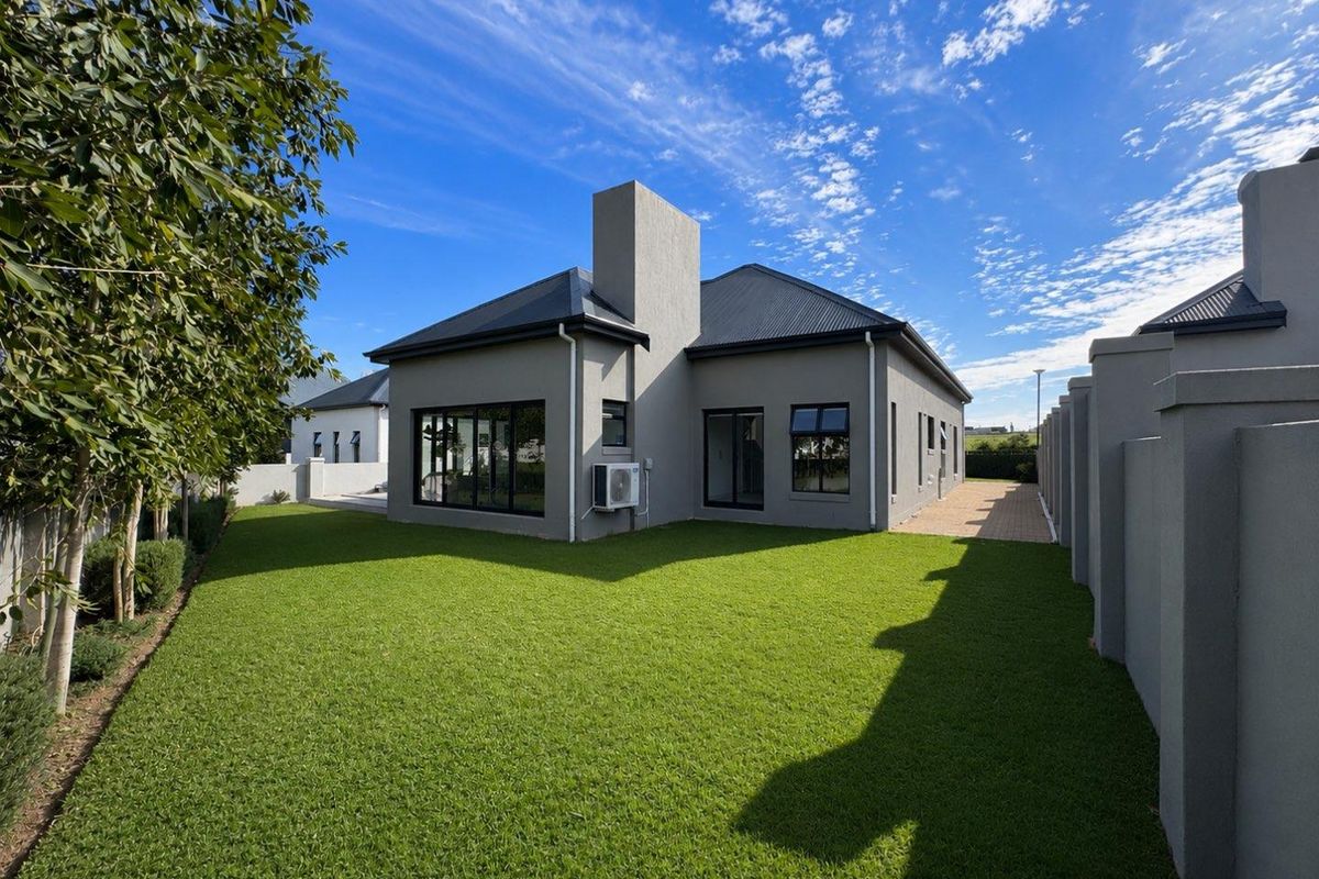 A well-maintained rear exterior view of the house featuring a manicured lawn, paved walkway, and modern grey facade with a prominent chimney.