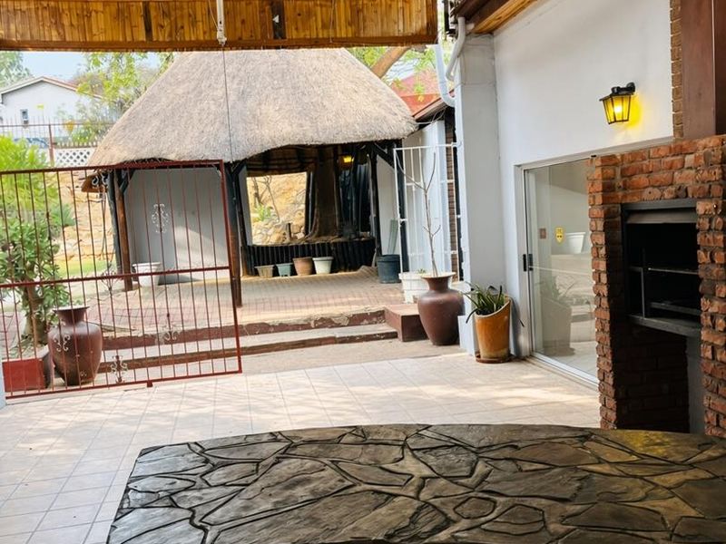 Covered outdoor patio featuring a built-in braai, stone-topped bar counter, and tiled flooring, overlooking a garden area with a thatched lapa.