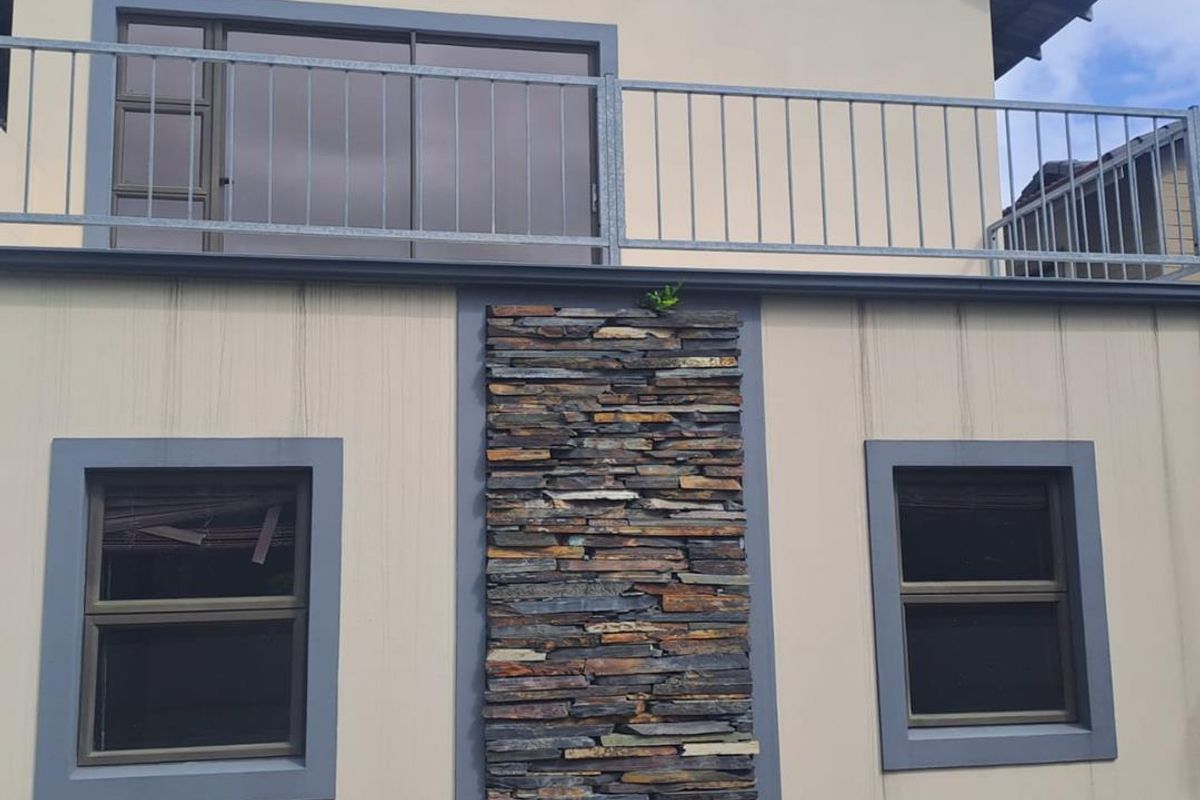 Rear exterior view of the property featuring a stone cladding accent wall, upper-level balcony with metal railing, and aluminium-framed windows.