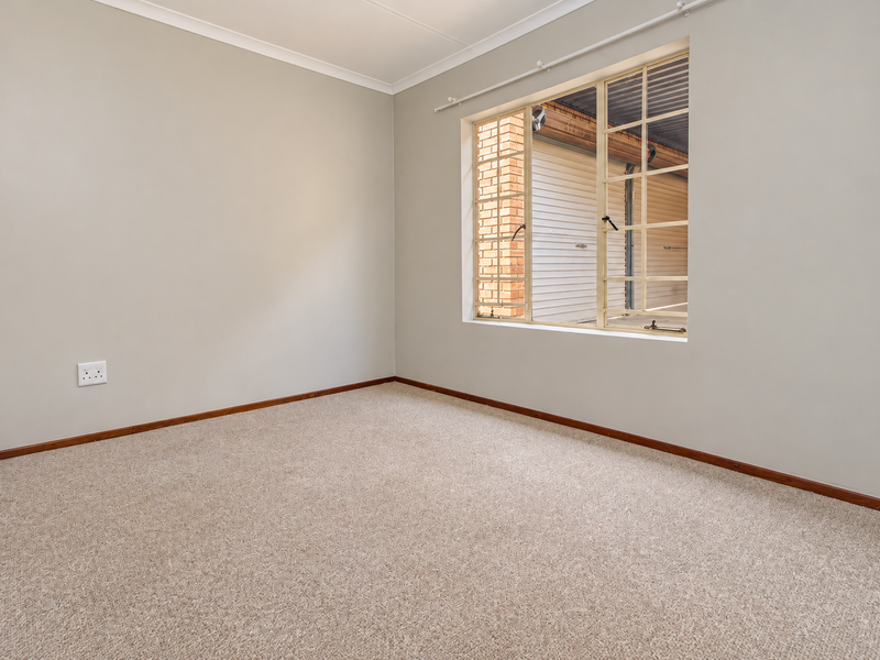 A clean, empty bedroom featuring neutral walls, carpeted flooring, and a window with security bars, offering a blank canvas for personalization.