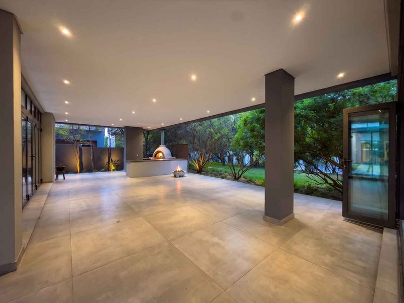 Spacious covered patio featuring large format floor tiles, recessed lighting, and a built-in pizza oven, seamlessly connecting to the garden through glass doors.