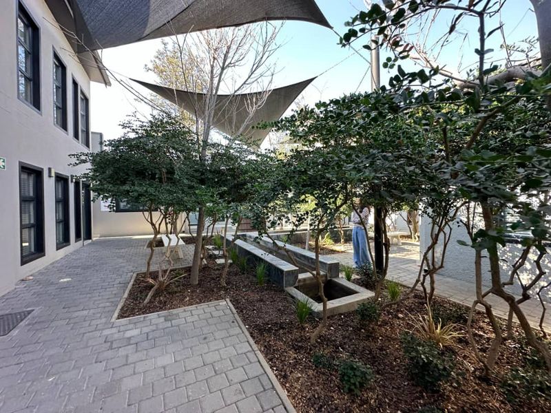 A well-maintained outdoor courtyard area featuring paved walkways, mature landscaping, raised garden beds, a water feature, and overhead shade sails for comfort.