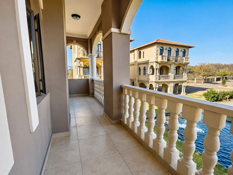 A spacious covered balcony featuring classic arched columns, a decorative balustrade, and neutral floor tiling, offering views of the surrounding estate.
