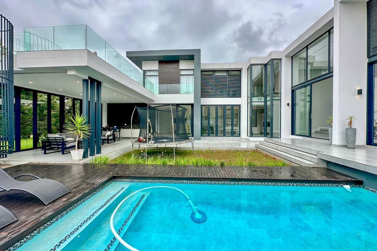 Modern architectural rear elevation featuring a swimming pool, timber decking, covered patio area, and extensive glass facade with upper-level balcony.