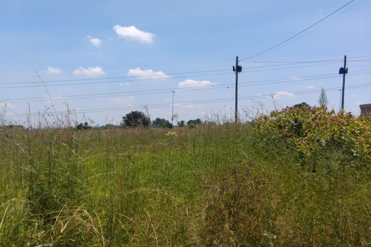 A view of a vacant residential plot featuring overgrown grass and visible utility infrastructure under a clear blue sky.