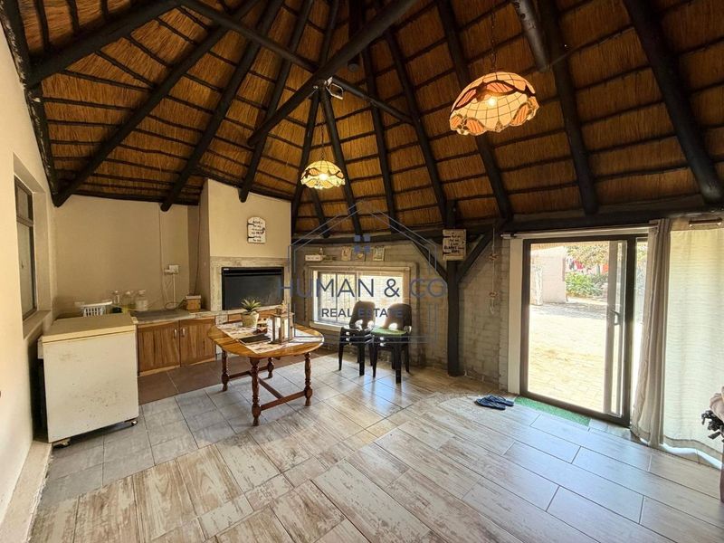 An enclosed patio area featuring a traditional thatched roof, exposed timber beams, built-in braai, and tiled flooring with direct access to the garden.