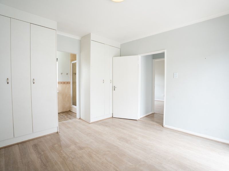 A bright, empty bedroom featuring light-toned laminate flooring, ample built-in cupboard space, and direct access to an en-suite bathroom.