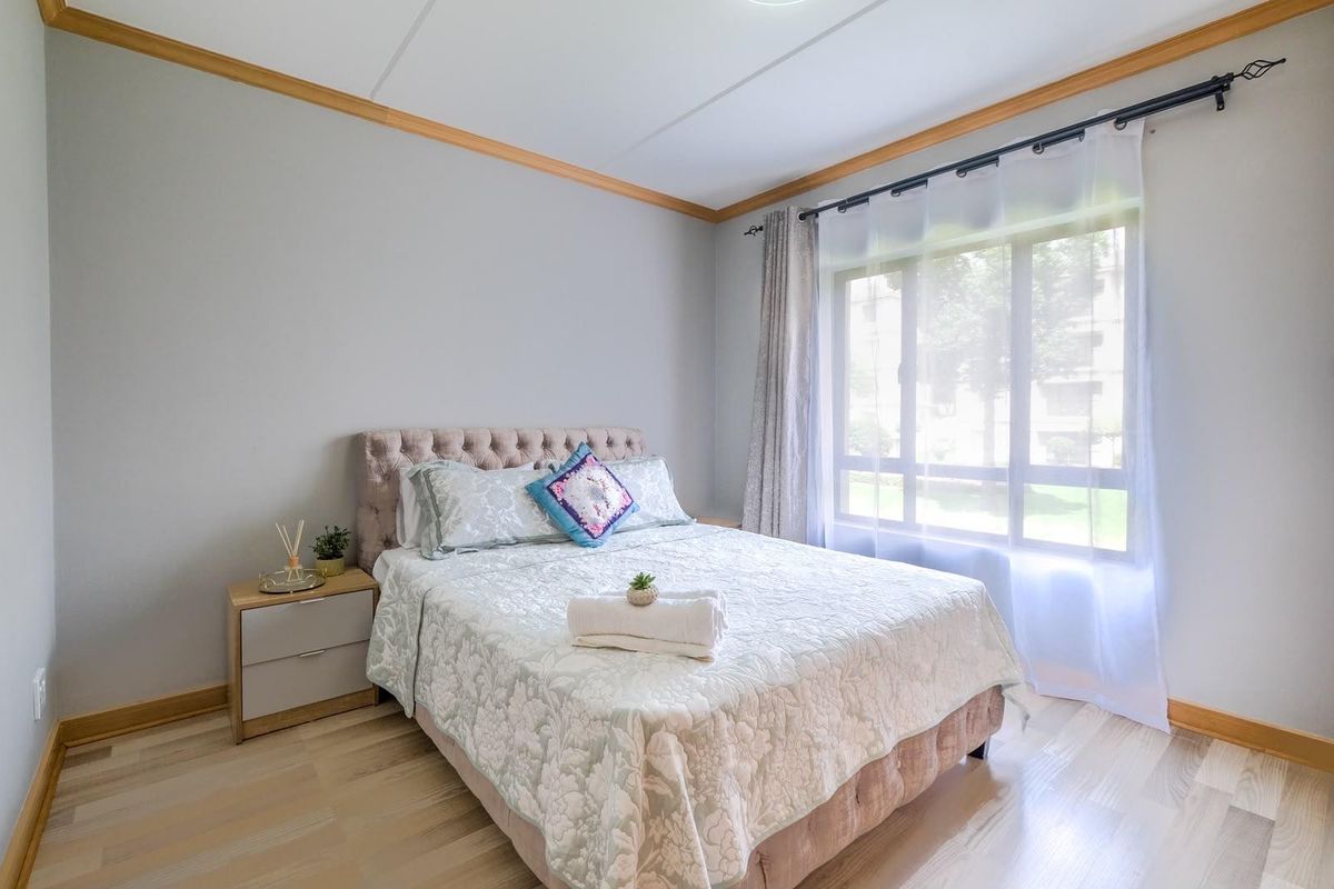 A well-lit bedroom featuring light-toned laminate flooring, timber crown molding, and baseboards, with a large window providing natural light.