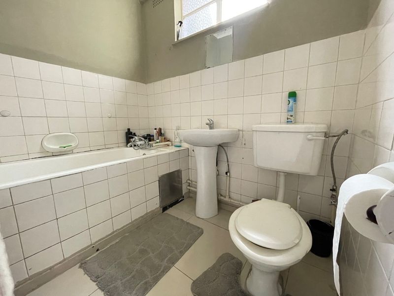 A dated bathroom featuring a standard bathtub, pedestal sink, and toilet with white wall tiling and visible plumbing fixtures.