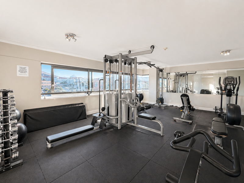 A well-maintained apartment gym featuring rubber flooring, mirrored walls, and large windows with natural light and urban views.