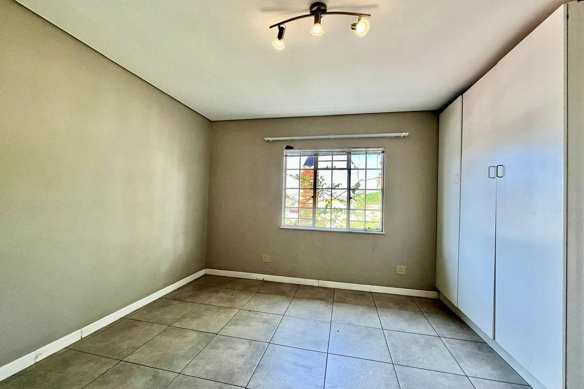 A bright, empty bedroom featuring neutral tiled flooring, built-in wardrobes, and a window with security bars.