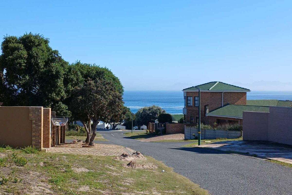 A street view of a residential area featuring a paved road, surrounding properties, and a clear view of the ocean in the distance.