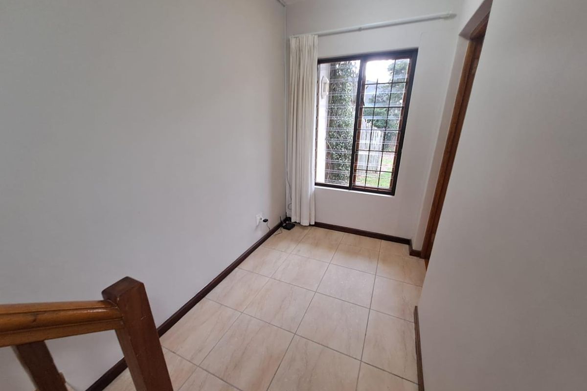 A bright, empty bedroom featuring tiled flooring, a window with security bars, and a wooden staircase railing.