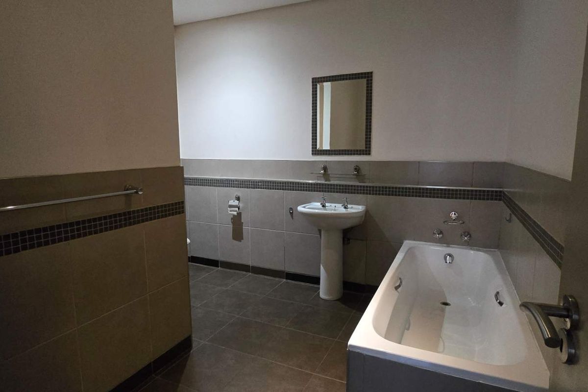 A clean, neutral-toned bathroom featuring a pedestal washbasin, a built-in bathtub, and tiled wall finishes.