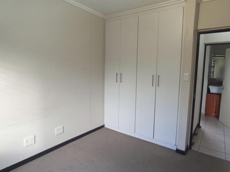 A clean, empty bedroom featuring neutral walls, carpeted flooring, and built-in white cupboards, with an adjacent view into a tiled bathroom.