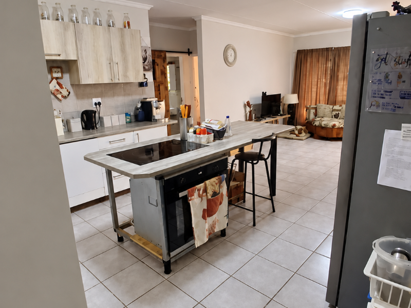 Open-plan living area featuring a central kitchen island with built-in hob and oven, tiled flooring, and neutral wall finishes throughout.