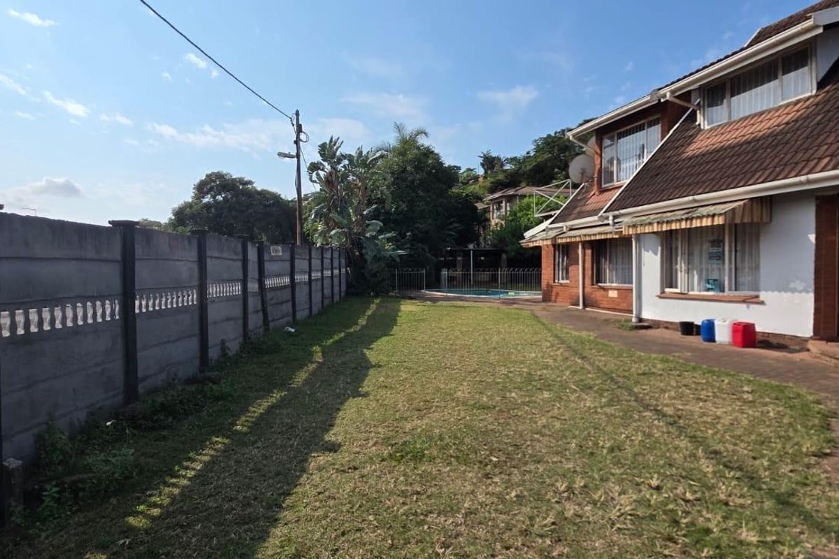 Rear exterior view of the property featuring a spacious lawn, paved walkway, swimming pool, and a secure precast concrete boundary wall.