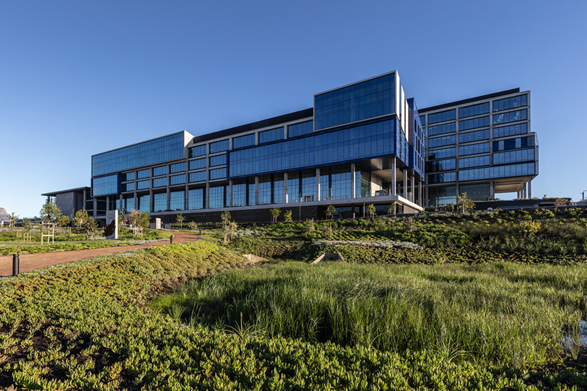A modern multi-story office building featuring a glass curtain wall facade, set within professionally landscaped grounds with a paved walkway.