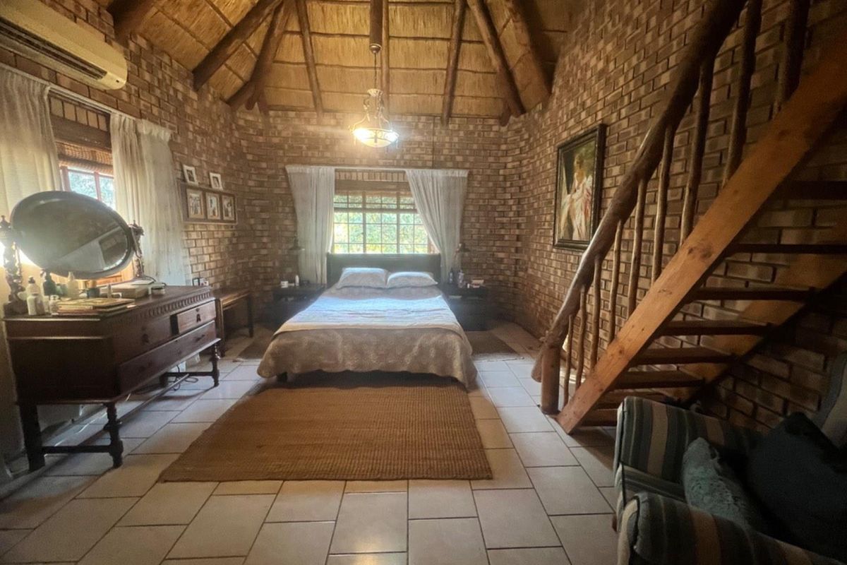 Spacious bedroom featuring exposed brick walls, a high thatched ceiling with timber beams, tiled flooring, and a wooden staircase leading to an upper level.