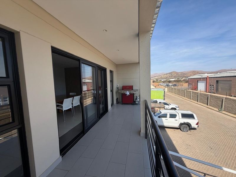 A spacious covered balcony featuring tiled flooring, modern sliding glass doors, and a metal safety railing, overlooking the surrounding business park area.