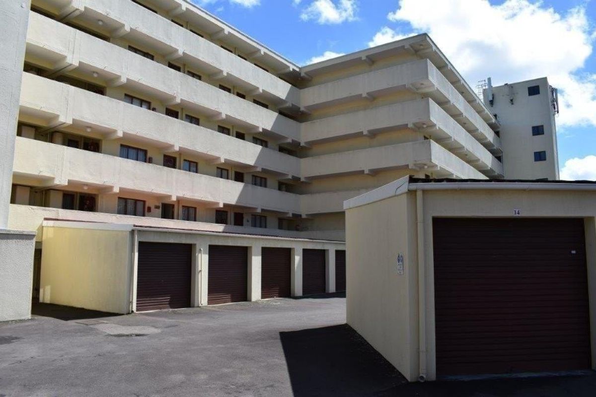 Exterior view of a multi-story apartment complex featuring a series of ground-level roller shutter garages and concrete balcony levels.